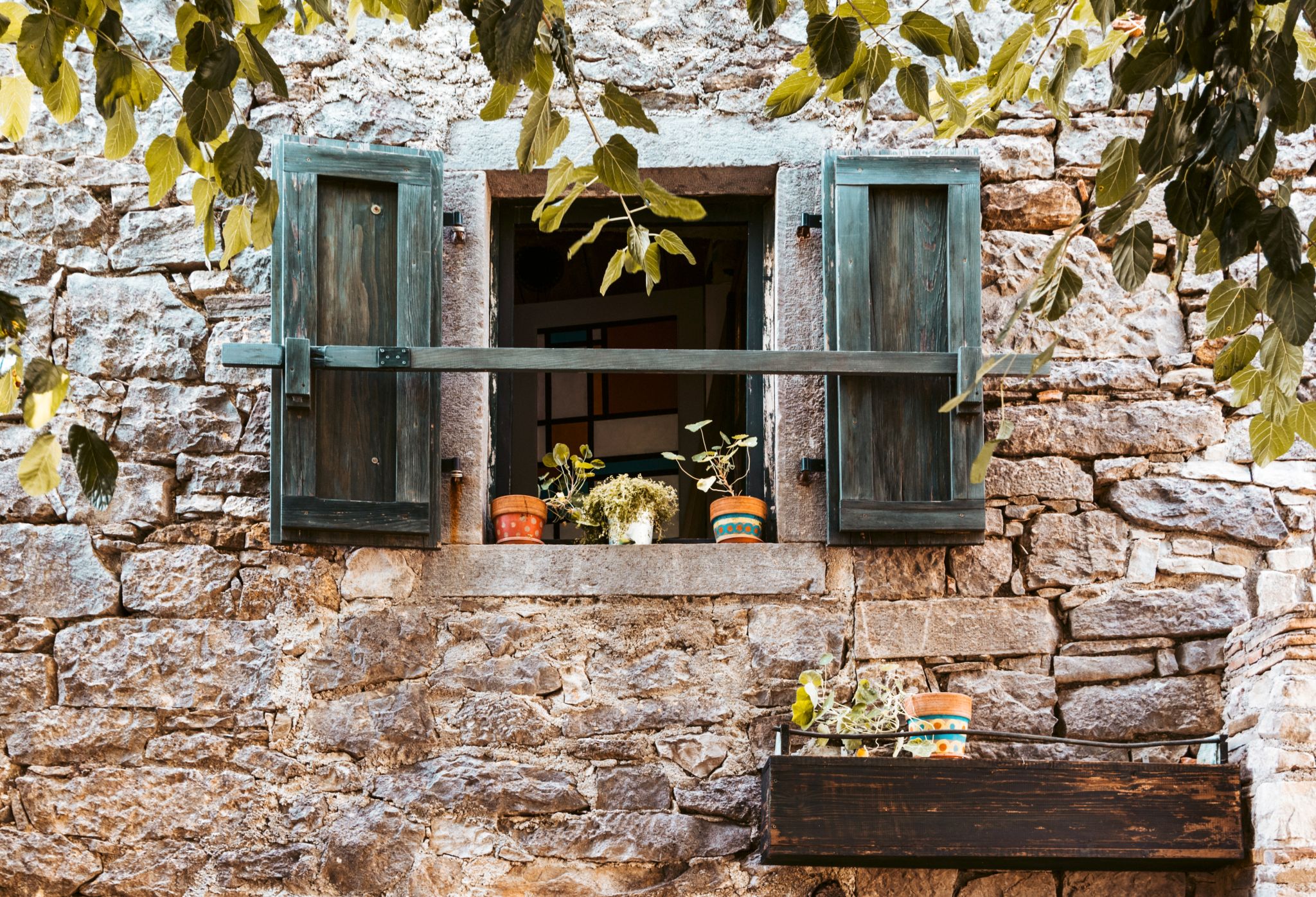 Window of a rural stone house with potted flowers on the window sill and wooden shutters.