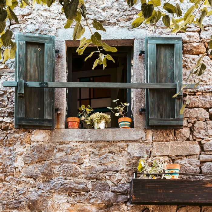 Window of a rural stone house with potted flowers on the window sill and wooden shutters.