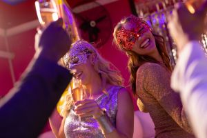 Zwei glückliche Frauen tragen Masken, tanzen und trinken Champagner auf einer Party in einem Nachtclub.