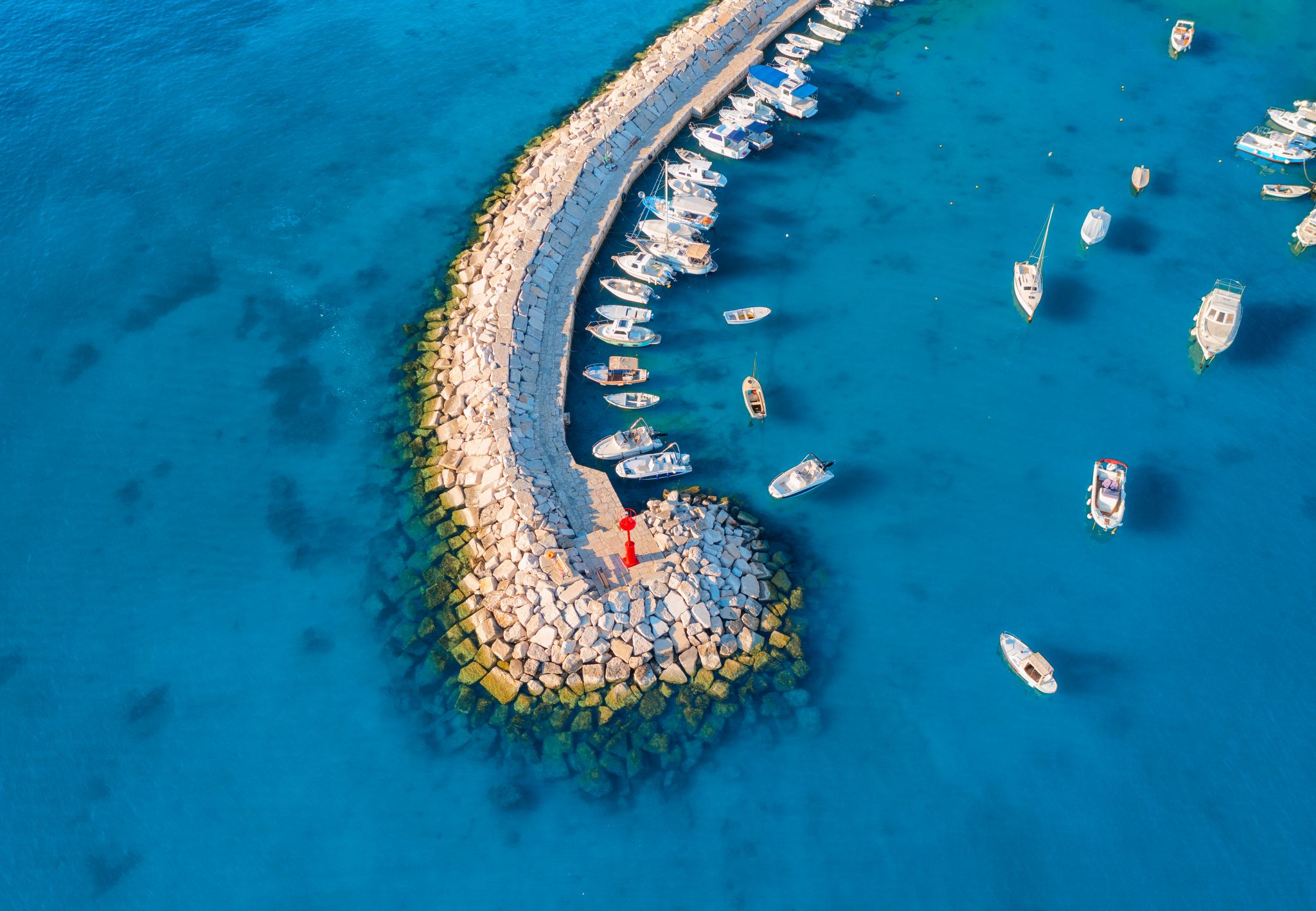 Aerial view of the vibrant harbor in Fažana, Croatia, with boats anchored along a curved stone pier extending into the clear blue Adriatic Sea.