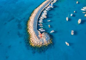 Aerial view of the vibrant harbor in Fažana, Croatia, with boats anchored along a curved stone pier extending into the clear blue Adriatic Sea.