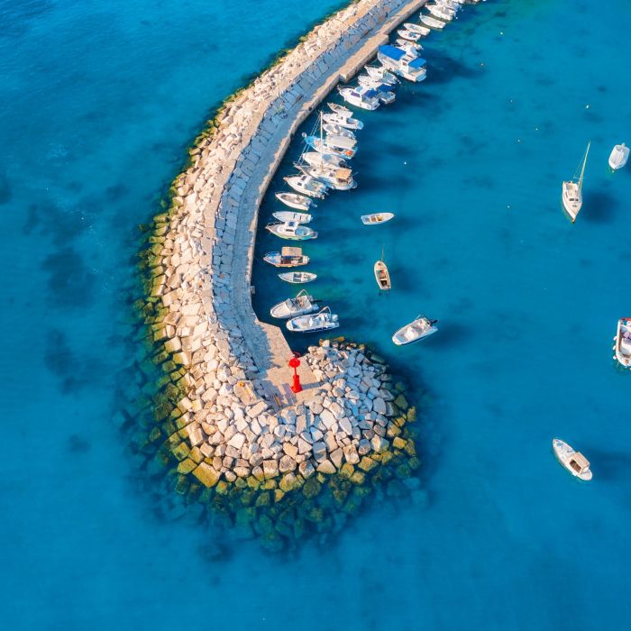 Aerial view of the vibrant harbor in Fažana, Croatia, with boats anchored along a curved stone pier extending into the clear blue Adriatic Sea.