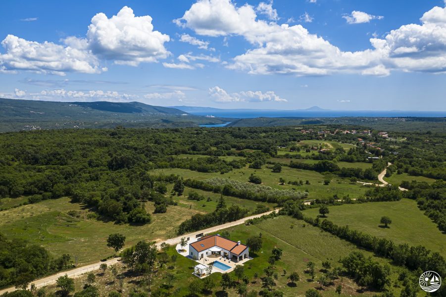 Zračni pogled na Villa Radola Residence okruženu bujnim zelenilom, s prostranim livadama, vijugavim stazama i udaljenim planinama ispod vedrog plavog neba.